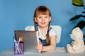 A girl 5-6 years old sitting at a table performs homework remote tasks on the tablet. Isolate on a...