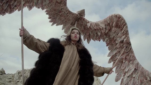 a young beautiful girl with long brown in a long dress and a fur cape depicts a bird flight against a blue sky, spreads its wings for flight, prepares for a mystical ceremony