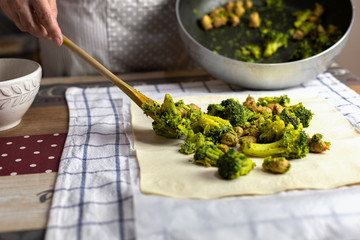 Female hand sprinkles broccoli with a wooden spoon on the surface of raw puff pastry.