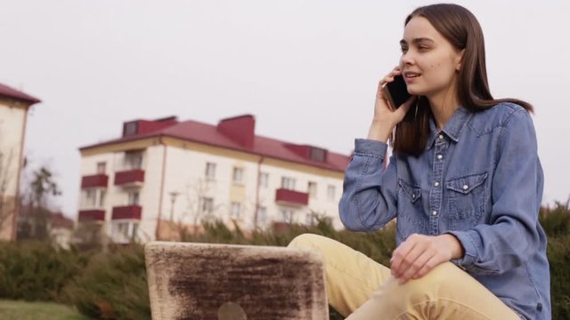 Young woman is talking on cellphone in street. Smiling female freelancer in casual wear speaking on smartphone and sitting on stone parapet in city during working break