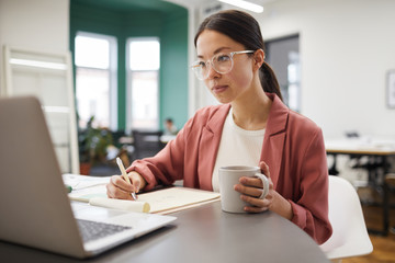 Young Asian businesswoman in eyeglasses looking at monitor of laptop drinking coffee and making notes at her workplace at office