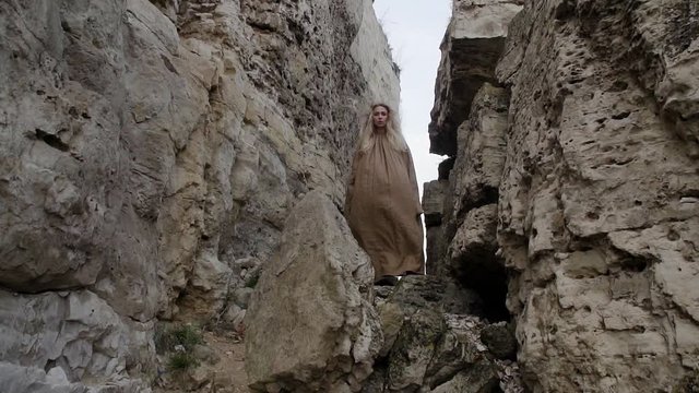young beautiful blonde girl is standing in a gorge of rocks in the mountains in a long dress, her hair and dress are developing in the wind