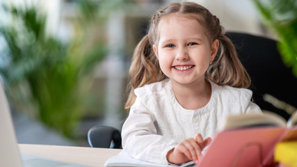 Cute laughing girl sitting at a table with a textbook. e-learning concepts