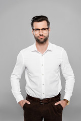 bearded businessman in glasses standing with hands in pockets isolated on grey