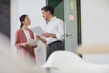 Two colleagues holding documents and talking to each other they discussing business documents while standing in the corridor at office