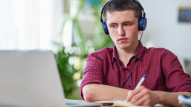 Brooding active teen guy with headphones listens to lecture, does in notebook
