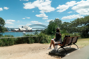 Woman with Sydney Opera House & Harbour Bridge. Tourist looking at attraction, with river water. Blue sky tourism shot. Boats on river. Famous landmark.