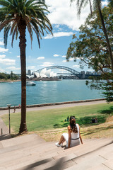 Woman with Sydney Opera House & Harbour Bridge. Tourist looking at attraction, with river water. Blue sky tourism shot. Boats on river. Famous landmark.
