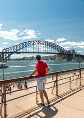 Man at Iconic Sydney Harbor Bridge over river water. Cityscape with buildings in CBD area. Tourist attraction in Australia. People at busy landmark. Black steel construction.