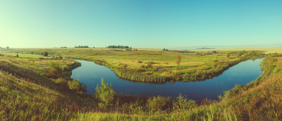 Obraz premium Sunny panoramic view of blue river and green meadows with trees on background.Retro and vintage colors. 
