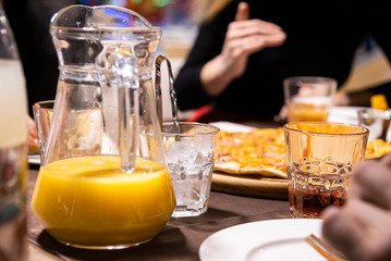 Close up of decanter with oranfe juice on the table in restaurant.  Friends party in restaurant. People in restaurant around the table. People's hands in restaurant