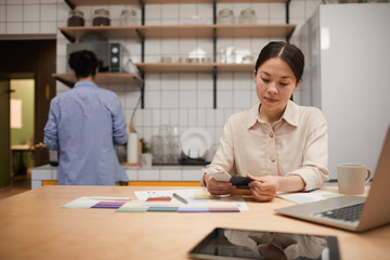 Serious young businesswoman sitting at the table with laptop and working with colored patterns in the kitchen at office