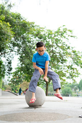 Outdoor portrait of a cute Malaysian little boy trying to jump from a concrete ball at the park.