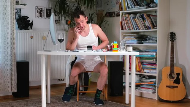 A man in shorts is dressing a shirt at a home office table. Children play under the table. Adapting a business to a home office, quarantine virus infection COVID 19.