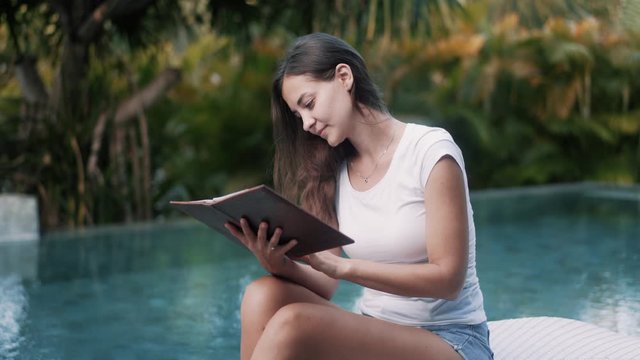 Young woman in summer clothes sits on sunbed and reads book near pool