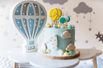 Children's first birthday cake, blue cake with clouds, meringue and balloons on the background of the children's room lights