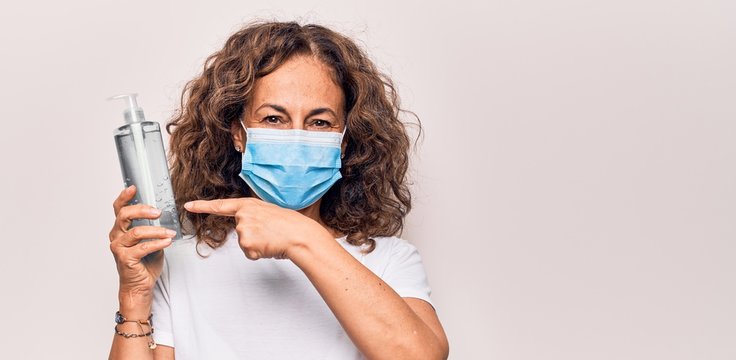 Middle age woman wearing coronavirus protection mask holding hand sanitizer bottle smiling happy pointing with hand and finger