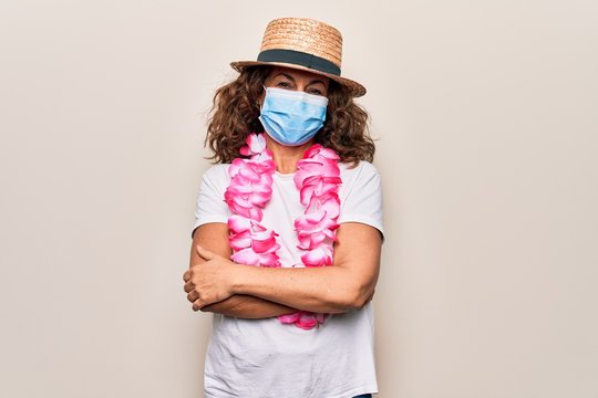 Middle age tourist woman wearing coronavirus protection mask and summer hat on vacation happy face smiling with crossed arms looking at the camera. Positive person.