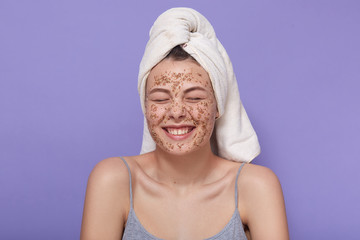 Studio shot of young attractive lady applies brown face scrub, looks happy, stands laughing with...