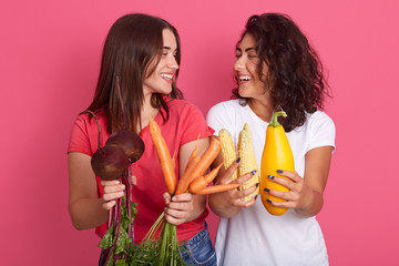 Indoor shot of two happy smiling women looking at each other, holding vegetables in hands:...