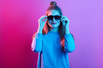 Closeup portrait of teenager female wearing casual sweater, posing with hair bun, woman keeping hands on sunglasses' frame, looking straight ahead, posing isolated over pink neon background.