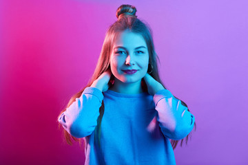 Fototapeta premium Indoor shot of attractive female wearing casual sweater, looking directly at camera, keeping both hands on neck, posing with hair bun isolated over pink neon background, lady has satisfied expression.