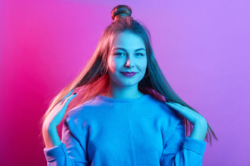 Studio shot of young adorable woman with long hair and bun, keeping hands near shoulders, touching her hair, looks at camera, standing against pink neon wall. People, teenager, good mood concept.,