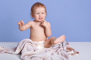 Closeup portrait of little child sitting on towel, calling mother, biting finger, looks unhappy,...