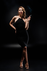 Fototapeta na wymiar full length view of sexy woman in elegant dress holding cigarette while posing on black