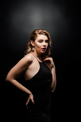 sexy, elegant woman looking at camera while posing with hand on hip on black background
