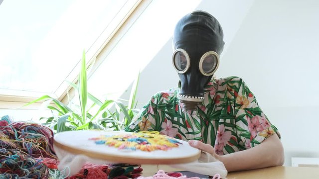 girl in gas mask at home during coronavirus pandemic COVID-19, sews with needle. embroidery hoop, fabric with colorful image. Stay home, quarantine leisure funny concept. Needlework, handicraft, hobby