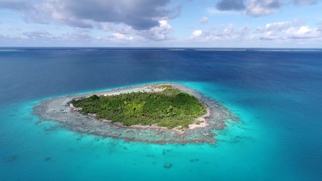 Aerial drone reveal shot of the bird island and turquoise waters of the Tikehau lagoon, French Polynesia. Palm trees and coconut trees fill the island. Blue sky with clouds fill the background