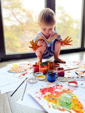 
A fair-skinned little child / infant, draws with colorful paints against the background of a large panoramic window at home.