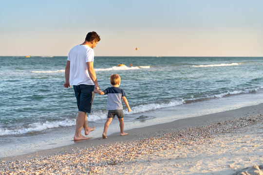 Young Caucasian dad with little son walk warm summer day along the sea coast. Summer family vacation concept. Friendship father and son.