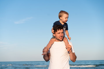 Young Caucasian dad with little son walk warm summer day along the sea coast. Summer family vacation concept. Friendship father and son.