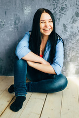 Cute long-haired brunette in a blue shirt and dark blue jeans is laughing, sitting on the wooden floor and leaning against the wall