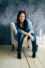 Cute long-haired brunette in a blue shirt and dark blue jeans is laughing while sitting in a chair and looking at the camera - 339912859