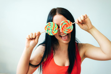 Close-up of a beautiful young girl hiding her eyes behind colorful lollipops on a stick, sticking out her tongue with a piercing - 339913042