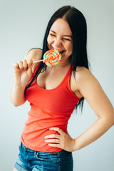 A cute cheerful girl in a bright orange t-shirt is nibbling colorful lollipop on stick - 339913086