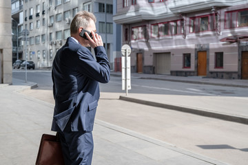 Fototapeta premium Alone middle-aged gray-haired businessman in a blue suit calling by phone standing on the empty city street.