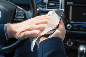 Middle age businessman in a blue suit cleaning mobile phone or gadget in a car using antivirus antibacterial wet wipe (napkin) for protect himself from bacteria and virus.