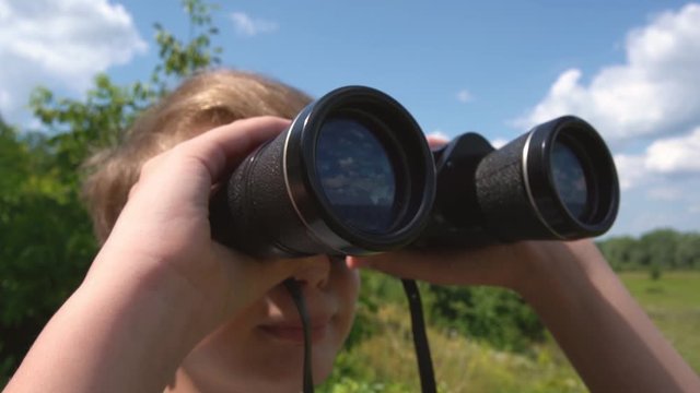 Closeup view video of cute white young kid looking through old black binoculars standing in sunny summer countryside landscape.
