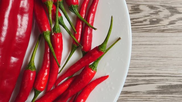 Top View Of Fresh Raw Red Chili Peppers And Ramiro Pepper With Water Drops Rotating On A White Plate On A Gray Wooden Background On The Left Side.