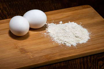 two white eggs on a wooden board and next to a handful of flour