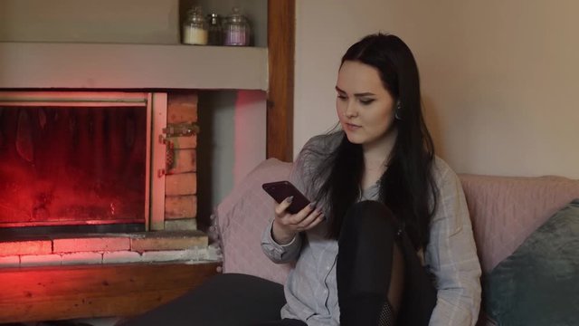The brunette is sitting at home and looking at the phone, the girl is chatting, playing games, smiling. The girl sits dma near the fireplace, all the time on the phone.