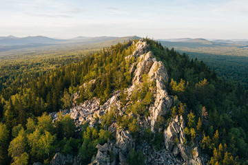 Rocky ridge among green forest at sunset, panoramic aerial view. Aleksandrovskaya Sopka - peak of Ural-Tau ridge. Ural wild nature landscape near Zlatoust in Chelyabinsk Oblast, Russia