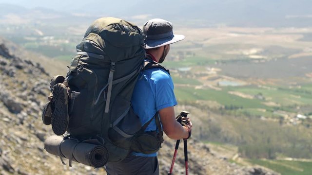 Mountain hiker with big backpack looks out over valley from mountainside on sunny day.