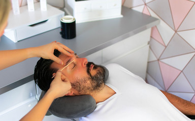 A beautician carries out an eyebrow wax on a relaxed man, removing unwanted hair and creating a flattering shape as part of a male grooming treatment and service in a fresh and modern beauty salon