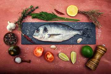 Dorado fish lies on a black stone cutting Board on beautiful pink concrete. Next are: lime, lemon, tomato, pepper, garlic, spices, salt and herbs.