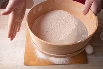 Large bowl, cutting board, sieve for sifting flour on the table. Female hands sifting flour in the home kitchen.